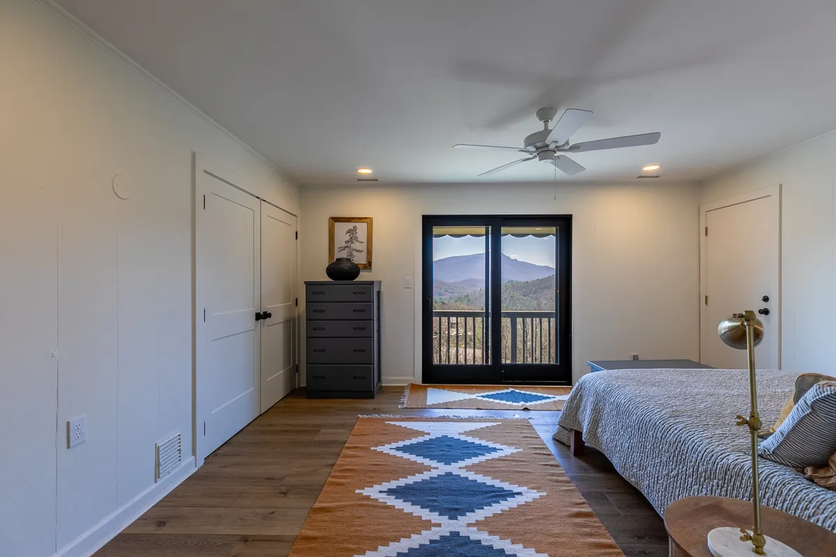 A peaceful bedroom with large windows framing stunning mountain views, a decorative rug, chest of drawers, bed with stylish bedding, and a soft lamp. The space feels calm, inviting, and perfect for relaxation or meditation.
