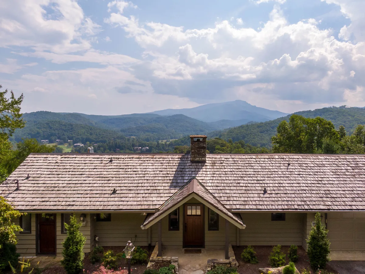 The image showcases the inviting facade of a beautifully renovated single-family mountain home, complete with a prominent stone chimney and a spacious front porch. The house is set against a breathtaking backdrop of Grandfather Mountain, with the rolling peaks visible in the distance. The porch looks perfect for relaxing, with ample space for gatherings, and the scene is bathed in tranquil sunset light, evoking a sense of peace and retreat.