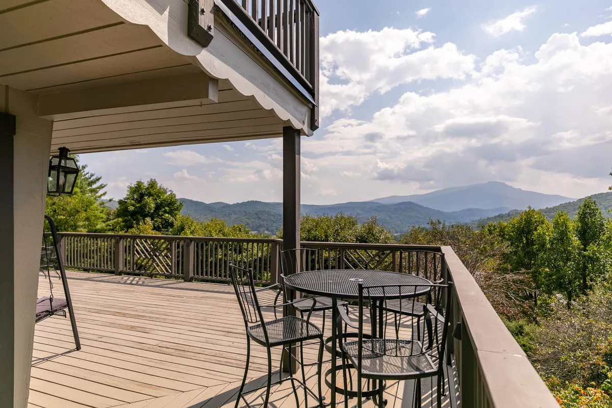 A wooden deck with railing overlooks a stunning mountain view, with a table and chairs positioned for outdoor dining or quiet reflection. The tranquil setting highlights the seamless transition from indoor to outdoor living, offering both group gathering spots and personal retreats.