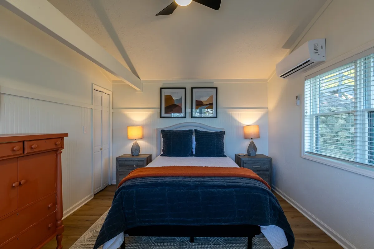 This image shows a tranquil bedroom with blue bedding, an orange throw blanket, nightstands with lamps, framed artwork, and a wooden dresser. The room feels restful, bright, and inviting—an ideal setting for peaceful sleep or a quiet morning.