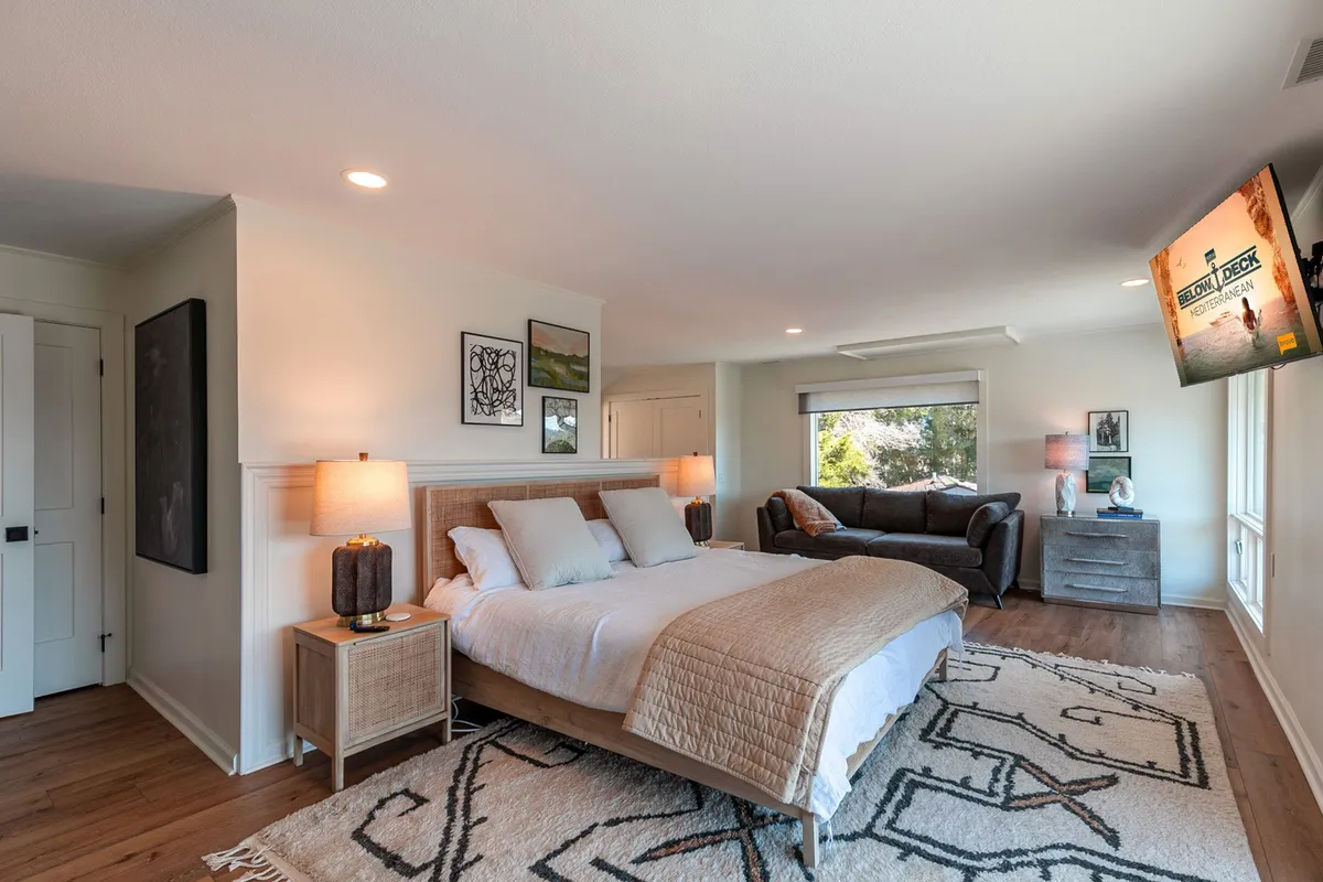 This image shows a comfortable, homey guest suite featuring a bed, a sofa, a table lamp, and a TV on the wall. The space is arranged to accommodate both relaxation and socializing, with ample natural light filtering in. The overall atmosphere is inviting and private—perfect for a restful stay.