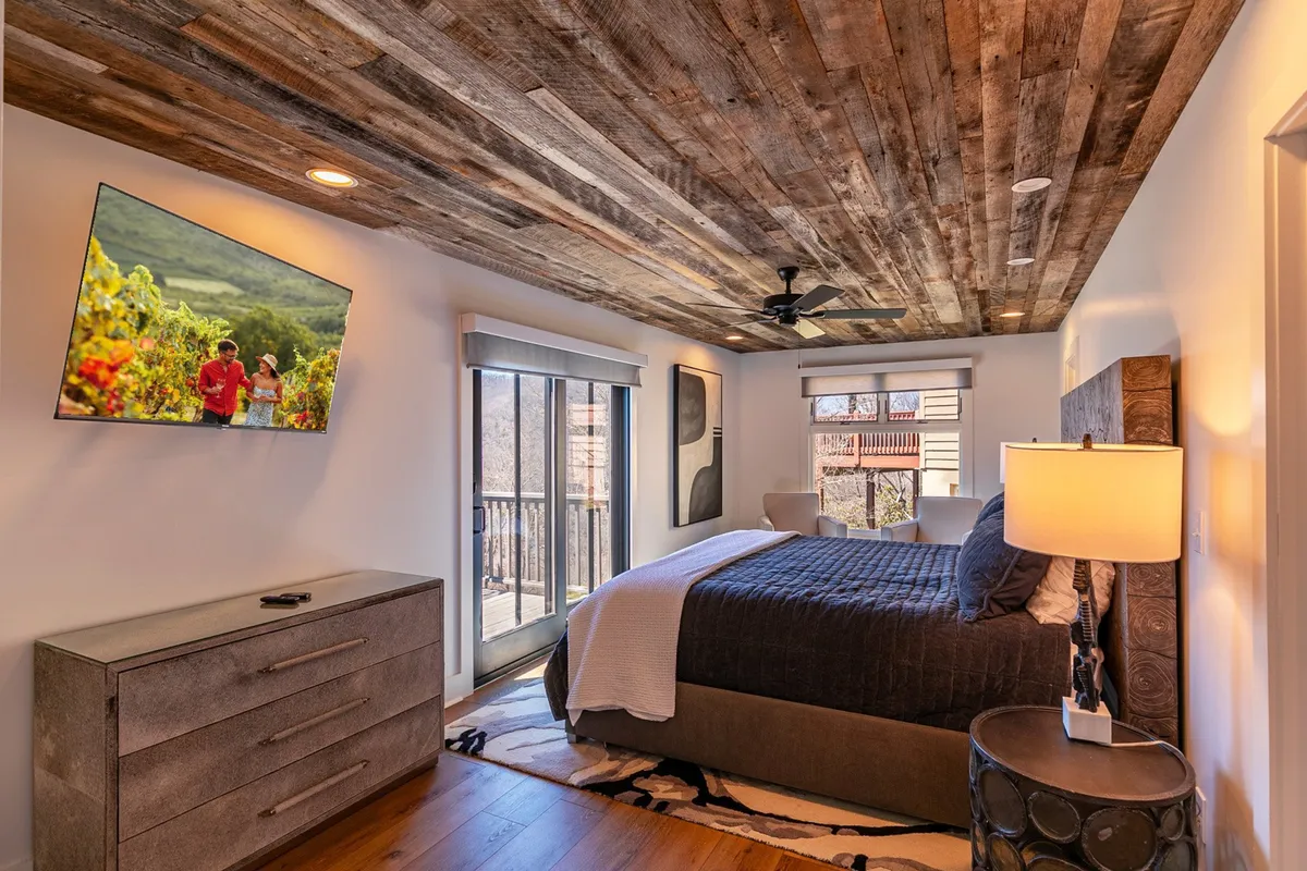 A tranquil suite with a cozy bed adorned in decorative bedding, a large TV displaying a scenic image, a modern dresser, stylish lamp, and a wooden ceiling. The room features a relaxing ambiance, and a balcony is visible through the window, emphasizing private outdoor access.