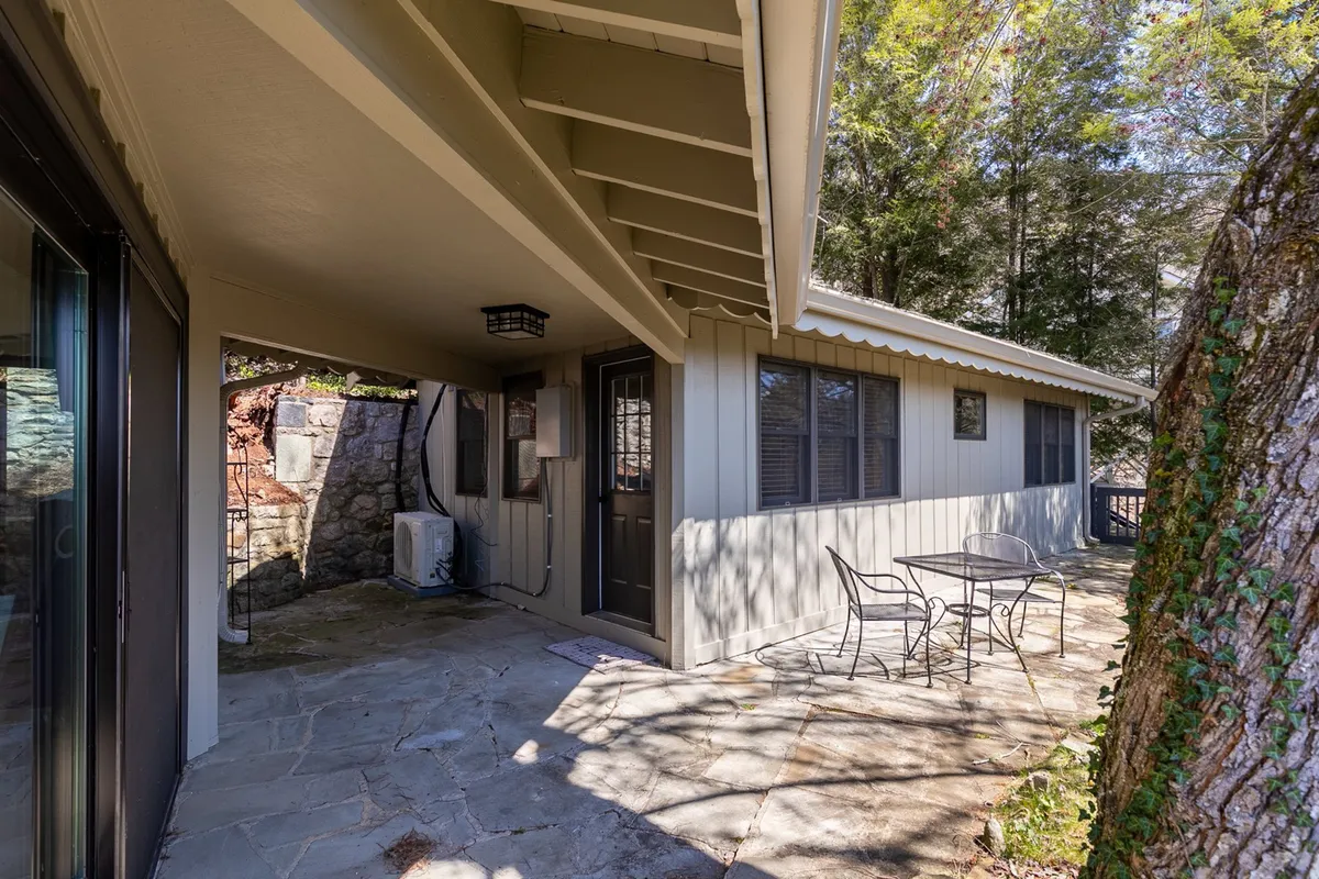 An intimate stone patio with a table and chairs sits beneath the canopy of surrounding trees, with the doorway to the house visible. The natural landscape envelops the space, ideal for outdoor dining or quiet moments away from the main living areas.