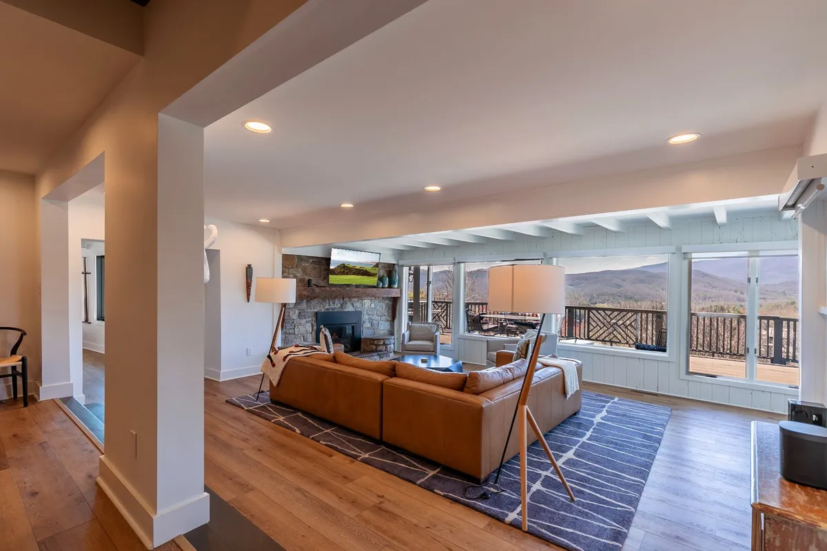 A cozy main living room featuring a leather sofa, stone fireplace, and expansive windows that frame stunning mountain views. The space is filled with warm natural light and includes a floor lamp and a plush rug, creating a welcoming environment for relaxation and gatherings.
