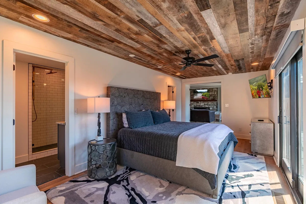 A serene bedroom featuring a king-sized bed with a textured headboard, warm wooden ceiling, large sliding glass doors that open to an outdoor space, and a decorative rug. Natural light fills the room, highlighting the inviting space and hinting at mountain views beyond.